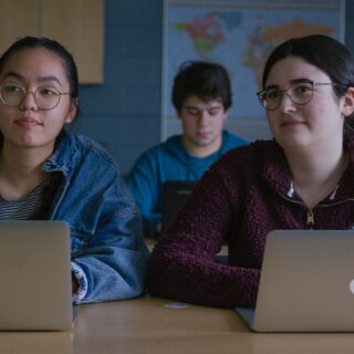 two-students-in-class-with-laptops-in-front-of-them-looking-toward-front